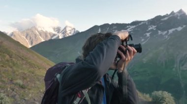 Adam kamerayı tutuyor, gözlerini kapıyor ve kameraya bakıp gülümsüyor.