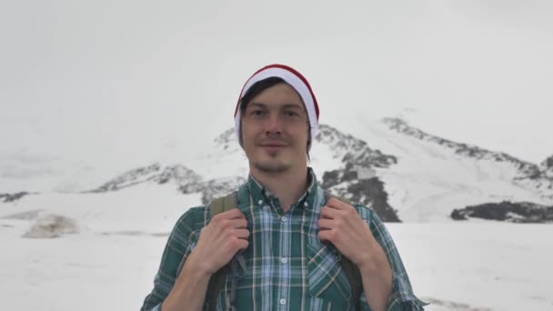 Молодий чоловік посміхається, приймаючи сніг на обличчі, стоячи на вершині пагорба — стокове відео