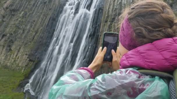 Lady håller telefonen och tar foto stående nära vattenfall — Stockvideo
