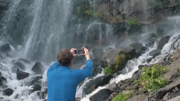 Homem detém telefone e faz vídeo em pé perto de cachoeira — Vídeo de Stock