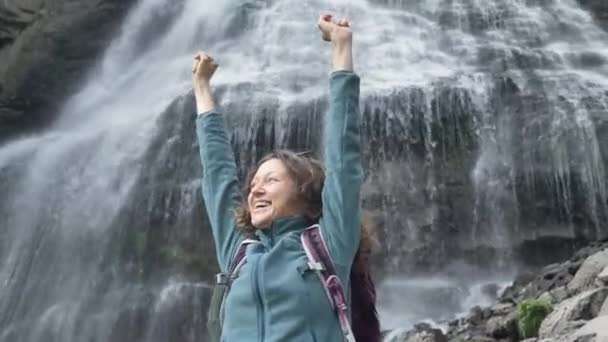 Счастливая леди с поднятыми руками радуется стоя у водопада — стоковое видео