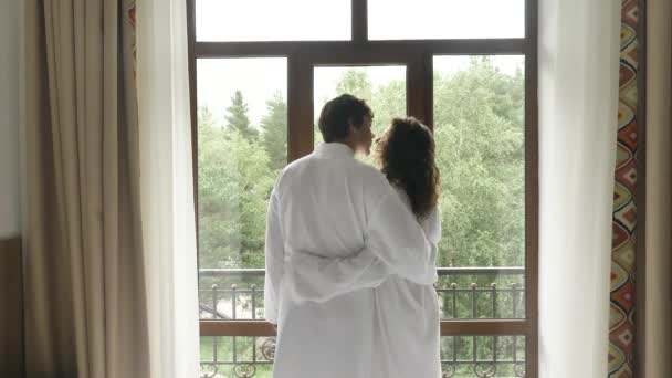 Jovem casal apaixonado em casacos brancos se aproximar da janela em um quarto de hotel, beijo, abraço e olhar para as vistas ao redor — Vídeo de Stock