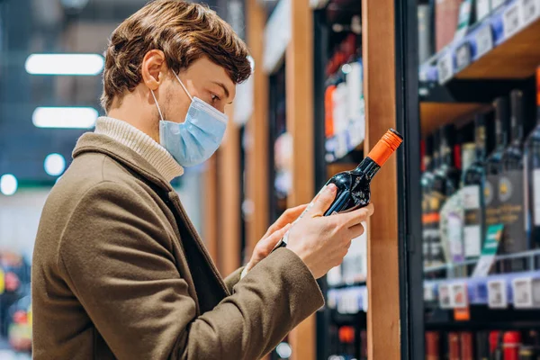 Успешный человек в маске читает этикетку на бутылке вина в супермаркете — стоковое фото