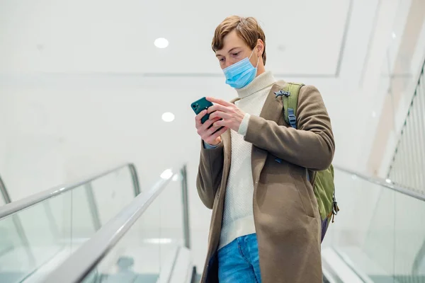 Человек в рюкзаке и маске едет на эскалаторе в торговый бизнес-центр и смотрит на телефон в руках — стоковое фото