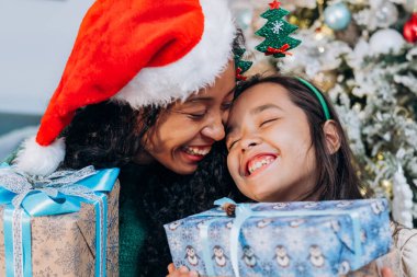 Anne ve kızı Noel hediyelerini tutuyorlar ve bir Noel ağacının arka planına gülüyorlar.