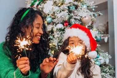Noel şapkalı bir anne ve küçük kız Bengal ışıkları tutuyorlar ve bir Noel ağacının arka planına gülüyorlar.