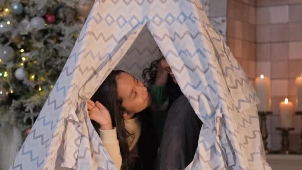 Mom and little daughter look out of the tent in the room and hold a garland in their hands against the background of a Christmas tree — Stock Video