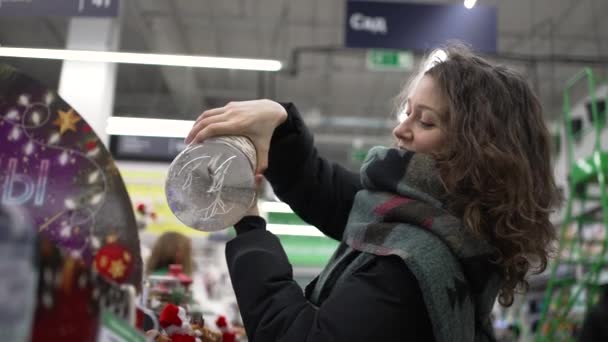 Hermosa mujer examina una bola de vidrio de Navidad en un supermercado — Vídeos de Stock