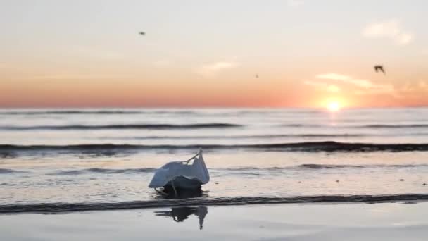 Tıbbi maske batan güneşe karşı denizin ıslak kumsalında yatıyor. — Stok video