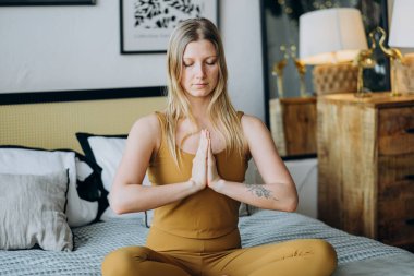 Sarışın kadın, gözleri kapalı yatakta meditasyon yapıyor.