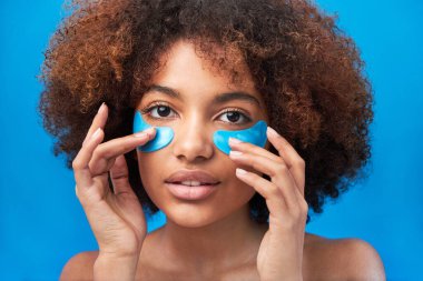 Kıvırcık saçlı kadın gözlerinin altında mavi bantlarla poz veriyor.