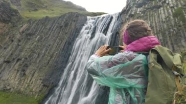 Kadın telefonu tutuyor ve şelalenin yanında fotoğraf çekiyor.
