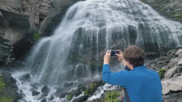 L'uomo tiene il telefono e fa video in piedi vicino alla cascata — Video Stock