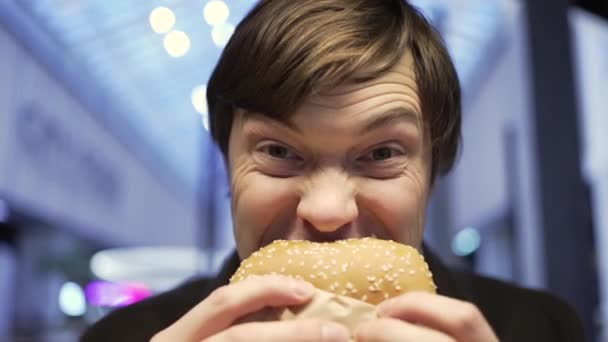 Portret van een jongeman die een hamburger bijt en naar de camera kijkt, close-up — Stockvideo
