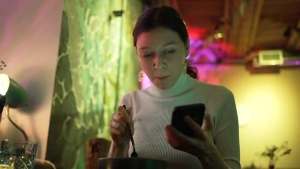 Ung vacker kvinna äter i en restaurang och tittar på en mobiltelefon mot en neon bakgrund — Stockvideo