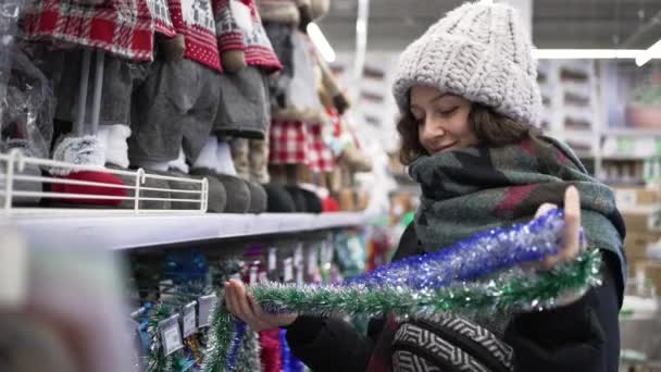 A young woman in a black down jacket in a supermarket chooses Christmas decorations — Video Stock