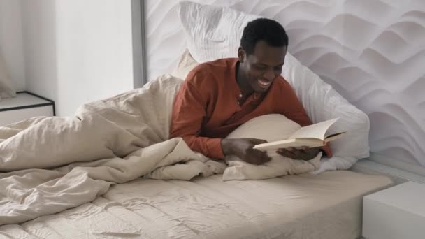 Happy African-American man in pajama reads book lying in bed — Stockvideo