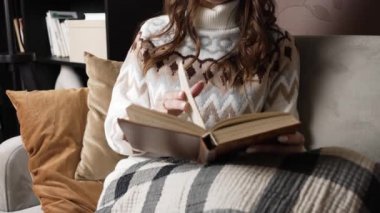 Woman covered with blanket sits on sofa and reads book
