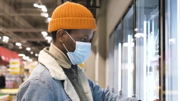 Young African-American man in a medical mask is shopping in a supermarket, goes to the refrigerator and chooses milk — Vídeo de Stock