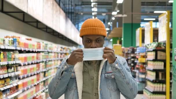 Portrait of an African-American man putting on a medical mask in a supermarket and giving a thumbs-up at the background of rows of groceries — 비디오