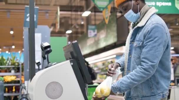 African-American male customer wearing a medical mask punches through items at a self-service checkout at a grocery supermarket — Wideo stockowe