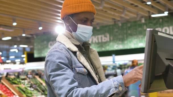 African-American man wearing a medical protective mask weighs fresh fruit on an electronic scale at a supermarket — стоковое видео