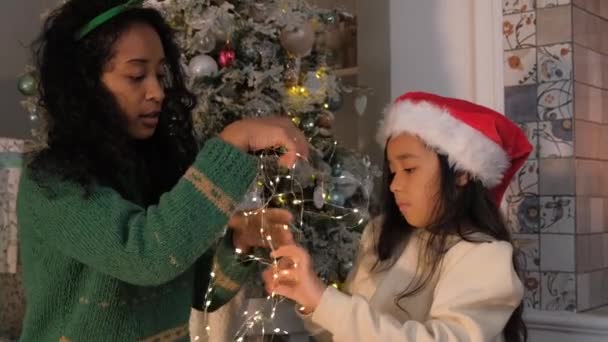 Daughter wraps mother with light garland by Christmas tree — Stockvideo