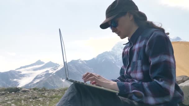 Леді сидить на вершині пагорба і типами на ноутбуку проти гір. — стокове відео