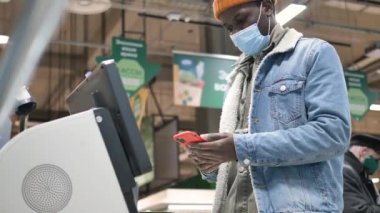 Afro- American guy types on smartphone at grey self-checkout