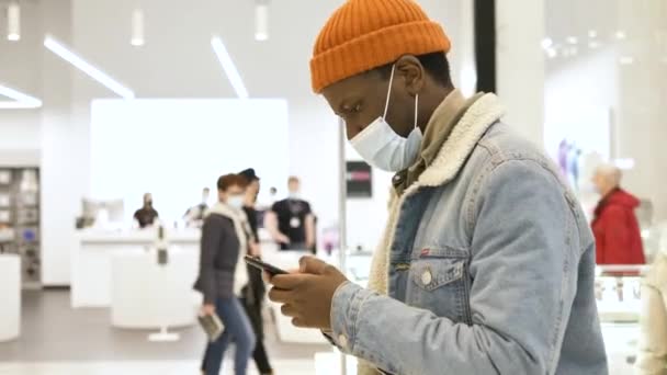 Concentrated black shopper in face mask types on smartphone — стоковое видео