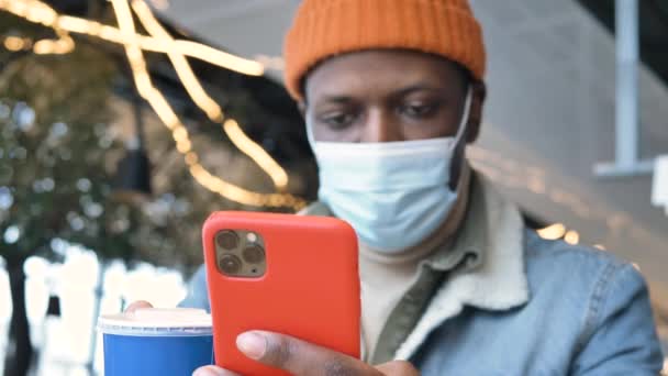 Concentrated black guy in mask holds smartphone and types — Vídeo de Stock