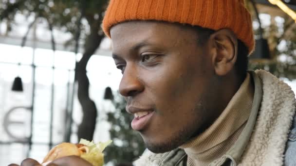 Black guy in jacket and hat tastes delicious burger in cafe — Wideo stockowe