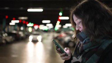 Elinde cep telefonu olan genç bir kadının portresi alışveriş merkezinin otoparkında yürüyor, ekrana bakıyor ve gülümsüyor.