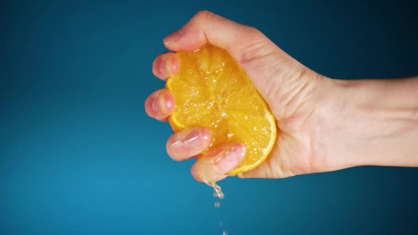 Жінки рука стискає половину апельсина, а сік повільно тече вниз пальцями на синьому фоні — стокове відео