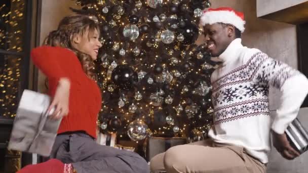 African-American couple simultaneously gives each other Christmas gifts on the background of a festive fir tree — Stockvideo