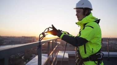 Industrial climber removes safety equipment from roof fence at sunset. Man in green overall finishes work on rooftop of skyscraper in evening