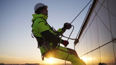 Industrial climber in working clothes and white helmet goes down wall of glass skyscraper. Worker in safety equipment climbs down building at sunset