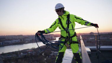 Industrial climber in a safety belt and a white helmet throws a rope while standing on the edge of a roof at sunset in slow motion