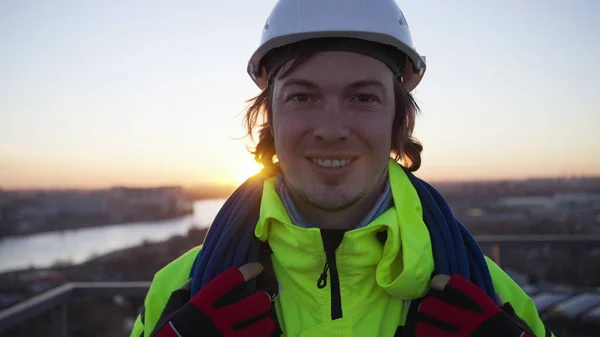 Young industrial climber in white helmet stands on rooftop at sunset. Climber with skein of rope on shoulders smiles looking in camera closeup