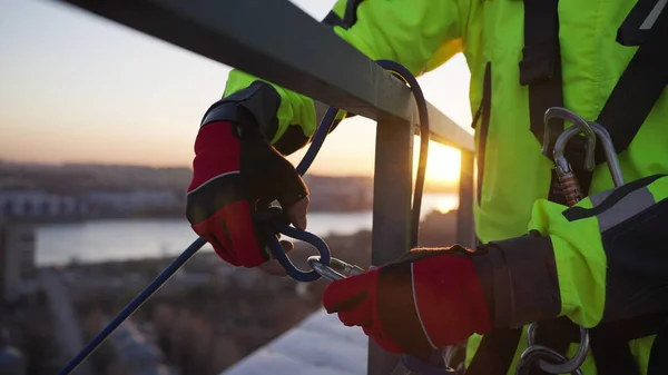 Man attaches rope with carabiner on roof steel fence. Industrial climber fastens safety equipment for climbing down building at sunset closeup