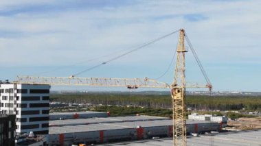High tower crane stands on construction site against industrial warehouses under cloudy sky. Building of new residential area in city aerial view