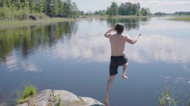 Young man with bare torso jumps into water from bank rock. Guy in black shorts swims in forest lake enjoying vacation far from city back view slow motion
