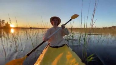 Young man does canoeing on calm forest lake with reeds. Guy in hat paddles kayak and enjoys relaxation. Person sails in canoe at sunset closeup