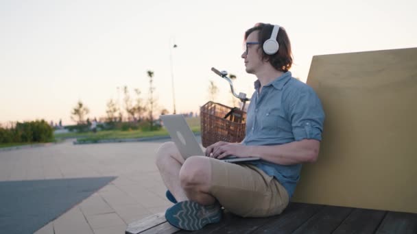 Freelancer Man Works Laptop Headphones Glasses Park Bench Sunset Summer — Stockvideo