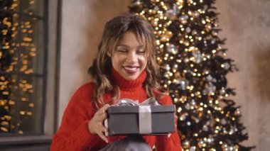 Young woman with curly brunette hair in red sweater opens box with present smiling and sitting near decorated Christmas tree