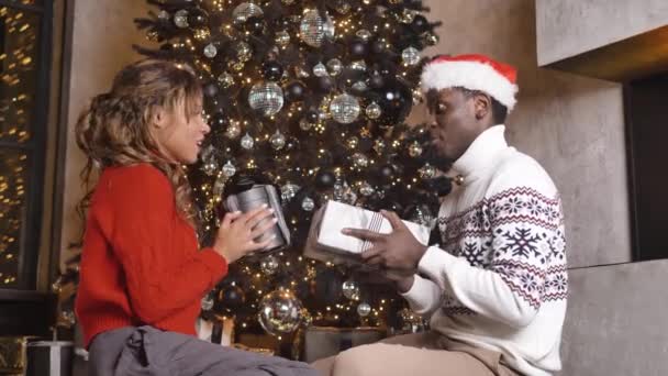 お祝いのセーターを着たアフリカ系アメリカ人のカップルは クリスマスプレゼントが飾りで飾られたクリスマスツリーの近くの床に座って開きます — ストック動画