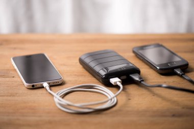 Siyah güç bankası USB kablosuna bağlı. Akıllı telefonu güç bankasıyla şarj ediyor. Güç bankası görüntüsüne odaklanmış sığ alan derinliği.