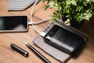 Siyah güç bankası USB kablosuna bağlı. Akıllı telefonu güç bankasıyla şarj ediyor. Güç bankası görüntüsüne odaklanmış sığ alan derinliği.