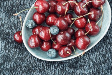 Koyu mavi örülü bir battaniye üzerinde olgun kırmızı kirazlarla dolu bir kase. Yukarıdan görüntüle.