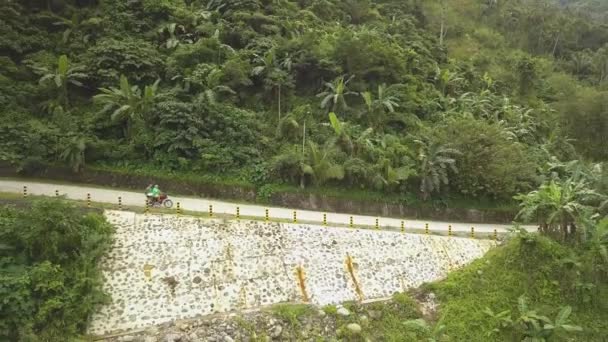 Hombre montando una motocicleta en una carretera con montañas en el fondo. Vista aérea de un joven montando un ciclomotor en un camino de montaña en el bosque — Vídeos de Stock